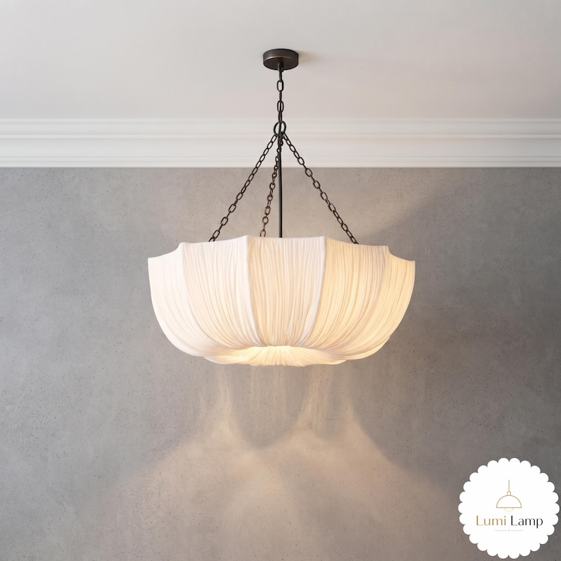 May include: A white fabric pendant light fixture with a pleated, bowl-shaped shade. The lamp is suspended by a dark metal rod and three thin chains connected to a circular ceiling canopy. The fabric emits a soft, diffused glow from within.