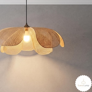 May include: A hanging pendant lamp featuring a woven tan rattan shade shaped like a large flower with multiple overlapping petals. A single light bulb is visible in the center, emitting a warm glow. The fixture hangs from a thin black cord.