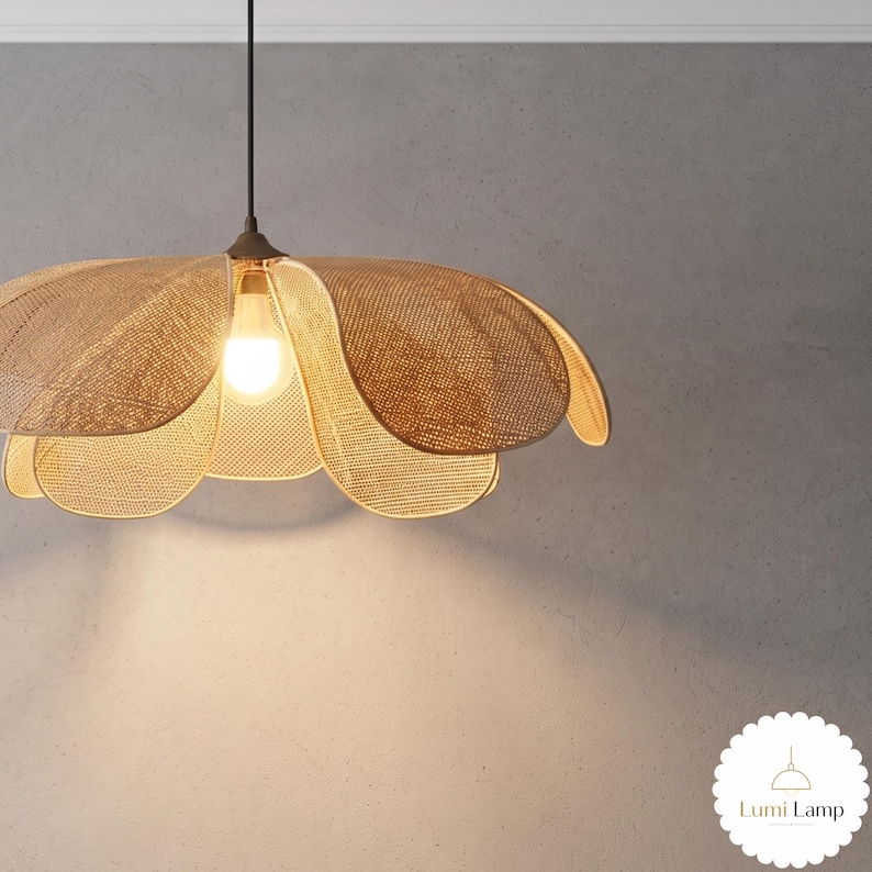 May include: A hanging pendant lamp featuring a woven tan rattan shade shaped like a large flower with multiple overlapping petals. A single light bulb is visible in the center, emitting a warm glow. The fixture hangs from a thin black cord.