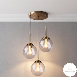 May include: A three-light pendant fixture featuring clear glass globes suspended from a circular brass ceiling canopy. Each globe houses a single light bulb with brass hardware and black cords. The text Lumi Lamp is visible in the bottom right corner.