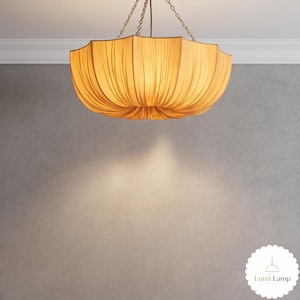 May include: A beige fabric pendant light with a scalloped, pleated shade suspended by three thin metal chains. The lamp emits a warm, downward glow. A small white circular logo in the corner features a line drawing of a lamp and the text Lumi Lamp.