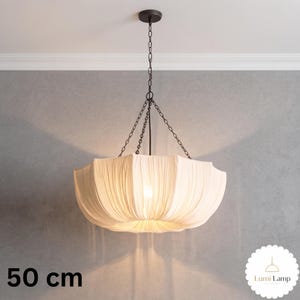 May include: A 50 cm pendant light featuring a wide, off-white fabric shade with vertical pleats. The shade is suspended by a dark metal chain assembly connected to a central rod and a circular ceiling canopy. The light emits a soft, warm glow.