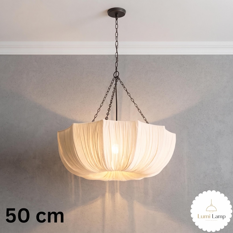 May include: A 50 cm pendant light featuring a wide, off-white fabric shade with vertical pleats. The shade is suspended by a dark metal chain assembly connected to a central rod and a circular ceiling canopy. The light emits a soft, warm glow.