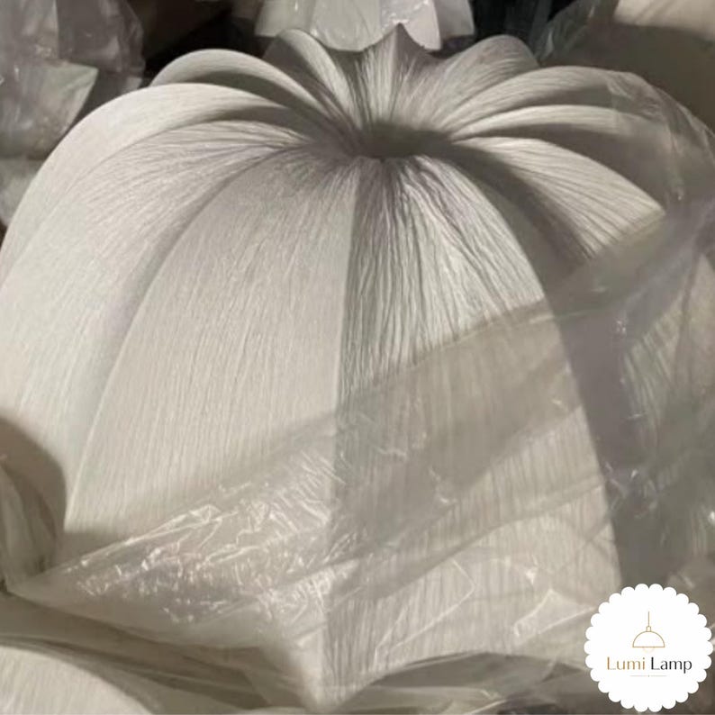 May include: A white, pleated, dome-shaped lamp shade with a textured, paper-like surface. The shade features a central opening at the top and is partially wrapped in clear plastic packaging. A small white circular logo reads Lumi Lamp.