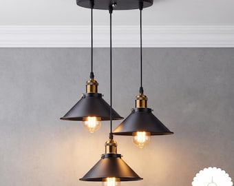 May include: A three-light pendant fixture featuring matte black conical metal shades suspended from a circular black ceiling canopy. Each shade includes a brass-toned socket housing a glowing Edison-style light bulb. The text Lumi Lamp is visible.