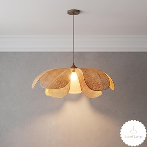 May include: A pendant light fixture featuring a woven tan rattan shade shaped like a blooming flower with multiple curved petals. The lamp hangs from a thin brown cord attached to a circular ceiling mount and emits a warm glow from a central bulb.
