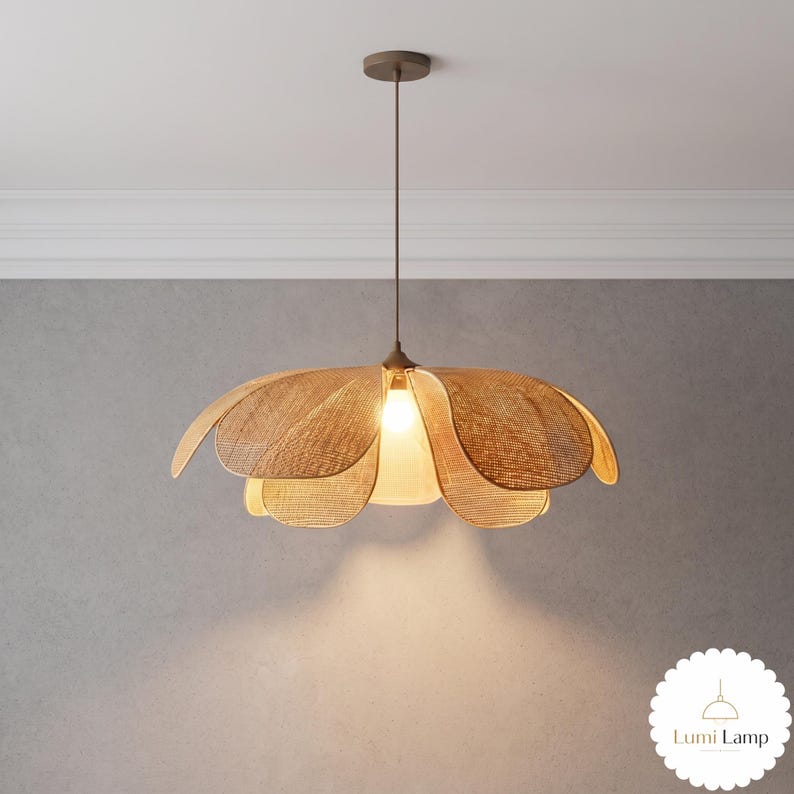 May include: A pendant light fixture featuring a woven tan rattan shade shaped like a blooming flower with multiple curved petals. The lamp hangs from a thin brown cord attached to a circular ceiling mount and emits a warm glow from a central bulb.