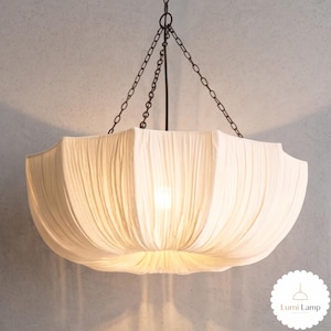 May include: A white fabric pendant light fixture with a gathered, pleated design suspended by dark metal chains. The shade has a wide, bowl-like shape with a soft, glowing light emanating from the center. A small logo reads Lumi Lamp.