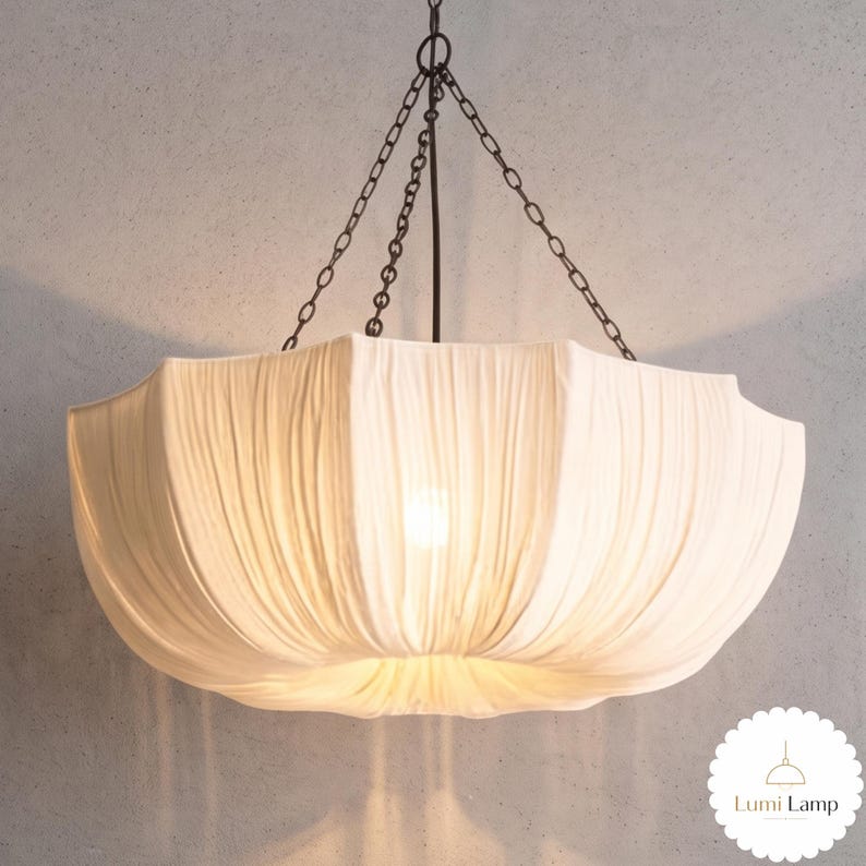 May include: A white fabric pendant light fixture with a gathered, pleated design suspended by dark metal chains. The shade has a wide, bowl-like shape with a soft, glowing light emanating from the center. A small logo reads Lumi Lamp.