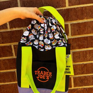 Może przedstawiać: Czarna torba płócienna z neonowo-żółtymi uchwytami i jasnofioletową podstawą. Torba posiada pomarańczowe, haftowane logo Trader Joe's oraz zapięcie na sznurek z czarnym wzorem przedstawiającym białe koty w kolorowych okularach w tęczowe wzory.