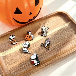 Peut inclure: Un ensemble de six broches en émail représentant le personnage Snoopy dans divers costumes d'Halloween, incluant une citrouille, un fantôme, une momie, une sorcière sur un balai et un vampire, présentées sur un plateau en bois clair.