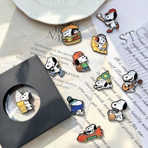 Könnte beinhalten: Eine Sammlung von Emaille-Anstecknadeln mit der Zeichentrickfigur Snoopy bei verschiedenen Aktivitäten, darunter Baseball spielen, einen Burger essen, Kopfhörer tragen, Basketball spielen, Gitarre spielen, Boba-Tee trinken und ein rotes Auto fahren.