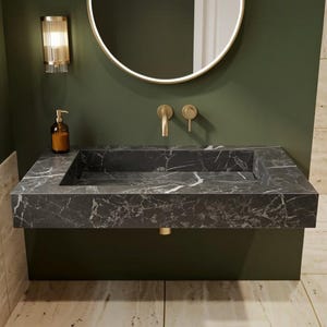 May include: A wall-mounted rectangular bathroom sink crafted from black marble with white veining. It features a gold-toned faucet and handle, a round backlit mirror, a glass soap dispenser, and a stack of white towels on a beige tiled surface.