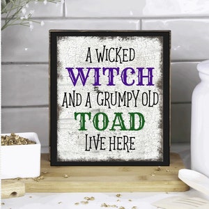 May include: A black framed sign with white distressed wood background. The sign reads "A Wicked Witch and a Grumpy Old Toad Live Here" in purple, black, and green lettering.