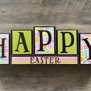 May include: Wooden block letters spelling "HAPPY EASTER" with colorful patterned paper on each block. The letters are black and the patterns are Easter themed.