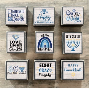 May include: Nine wooden blocks with white and gray wood grain designs. Each block features text and graphics related to Hanukkah, including phrases like "Happy Chanukkah," "Let's Get Lit," and "Eight Crazy Nights." The blocks have a distressed, rustic appearance.