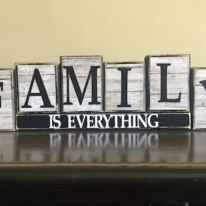 May include: Wooden block letters spelling "FAMILY" with a black and white woodgrain finish. The letters are arranged on a black surface with the words "IS EVERYTHING" written in white.