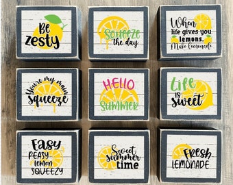May include: Nine wooden blocks with black frames and white backgrounds featuring lemon-themed sayings. The sayings include "Be Zesty", "Squeeze the Day", "When life gives you lemons, make lemonade", "You're my main squeeze", "Hello Summer", "Life is sweet", "Easy Peasy Lemon Squeezy", "Sweet Summer Time", and "Fresh Lemonade".