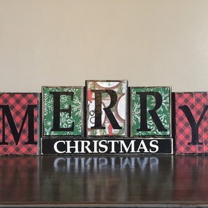 May include: Wooden block letters spelling "Merry Christmas" with red and green plaid patterns and a black background. The letters are arranged in a line on a dark brown surface.
