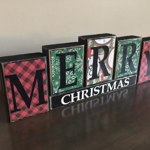 May include: Wooden block letters spelling "Merry Christmas" with a red and black plaid pattern on the "M" and "Y" blocks. The other letters have a green and white snowflake pattern.