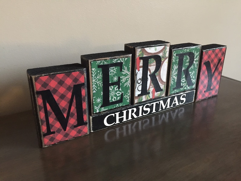 May include: Wooden block letters spelling "Merry Christmas" with a red and black plaid pattern on the "M" and "Y" blocks. The other letters have a green and white snowflake pattern.