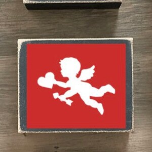 K&ouml;nnte beinhalten: Drei Holzschildchen mit Valentinstag-Motiv. Das oberste Schild ist rosa mit einem Tic-Tac-Toe-Spielfeld mit Herzen und X. Das mittlere Schild ist rot mit einer wei&szlig;en Silhouette von Amor. Das unterste Schild ist rosa mit dem Wort "LOVE" in schwarzen Buchstaben und einem rot-schwarzen karierten Herzen.