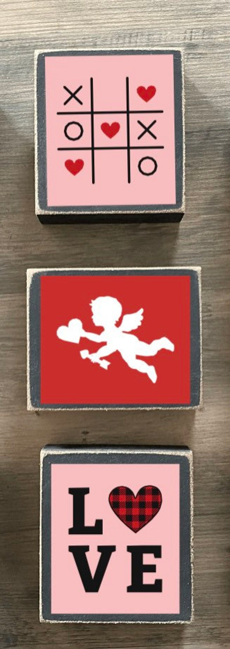 K&ouml;nnte beinhalten: Drei Holzschildchen mit Valentinstag-Motiv. Das oberste Schild ist rosa mit einem Tic-Tac-Toe-Spielfeld mit Herzen und X. Das mittlere Schild ist rot mit einer wei&szlig;en Silhouette von Amor. Das unterste Schild ist rosa mit dem Wort "LOVE" in schwarzen Buchstaben und einem rot-schwarzen karierten Herzen.