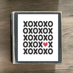 K&ouml;nnte beinhalten: Drei Holzschildchen mit schwarzen Rahmen und wei&szlig;em Text. Das oberste Schild sagt "Be Mine" mit einem rot-schwarzen karierten Herzen. Das mittlere Schild sagt "XOXO" in einem Raster wiederholt mit einem kleinen roten Herzen in der Mitte. Das unterste Schild sagt "Happy Valentine's Day" mit Pfeilen auf beiden Seiten des Textes.