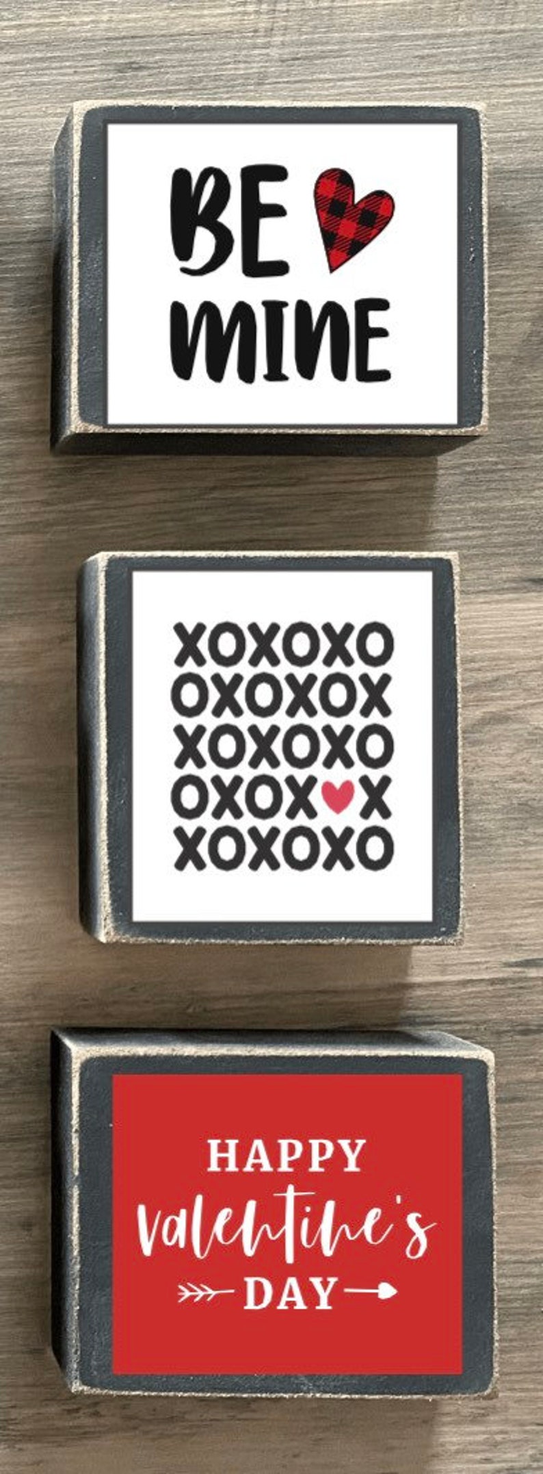 K&ouml;nnte beinhalten: Drei Holzschildchen mit schwarzen Rahmen und wei&szlig;em Text. Das oberste Schild sagt "Be Mine" mit einem rot-schwarzen karierten Herzen. Das mittlere Schild sagt "XOXO" in einem Raster wiederholt mit einem kleinen roten Herzen in der Mitte. Das unterste Schild sagt "Happy Valentine's Day" mit Pfeilen auf beiden Seiten des Textes.