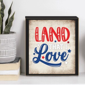 以下が含まれることがあります： 「LAND THAT I Love」の文字が赤と青で書かれ、星のアクセントが付いた額入りのプリント。プリントはヴィンテージ風で、くすんだ外観で、ニュートラルな背景に配置されています。フレームは黒です。