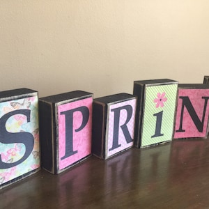 May include: Wooden block letters spelling "SPRING" with a pink, green, and white floral pattern. The letters are painted black and have a distressed finish.