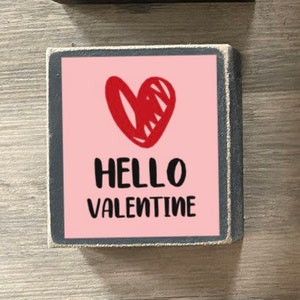 K&ouml;nnte beinhalten: Drei Holzschildchen mit Valentinstags-Botschaften. Das oberste Schild ist rot mit wei&szlig;em Text, der "Hugs and Kisses" lautet. Das mittlere Schild ist rosa mit einem roten Herzen und wei&szlig;em Text, der "Hello Valentine" lautet. Das unterste Schild ist wei&szlig; mit schwarzem Text, der "Will you be my Valentine?" lautet, mit einem roten Herzen und einem Pfeil.