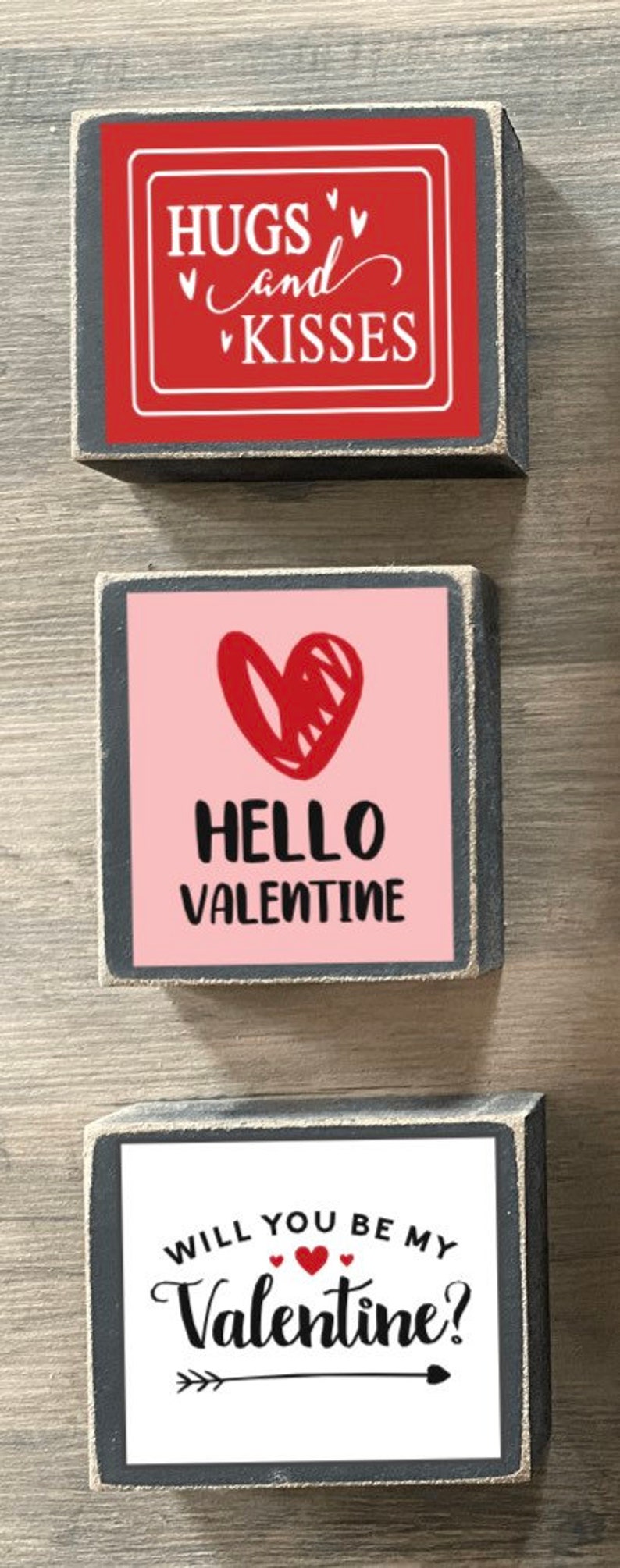 K&ouml;nnte beinhalten: Drei Holzschildchen mit Valentinstags-Botschaften. Das oberste Schild ist rot mit wei&szlig;em Text, der "Hugs and Kisses" lautet. Das mittlere Schild ist rosa mit einem roten Herzen und wei&szlig;em Text, der "Hello Valentine" lautet. Das unterste Schild ist wei&szlig; mit schwarzem Text, der "Will you be my Valentine?" lautet, mit einem roten Herzen und einem Pfeil.