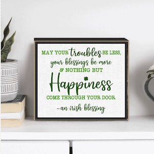 May include: A black framed sign with a white background and green text. The sign reads "MAY YOUR troubles BE LESS, your blessings be more & NOTHING BUT Happiness COME THROUGH YOUR DOOR. -an irish blessing".