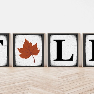 Op de afbeelding: Vier houten borden die het woord "FALL" spellen met een bruin esdoornblad in het midden. De borden hebben een witte achtergrond met zwarte letters en een verweerde look.