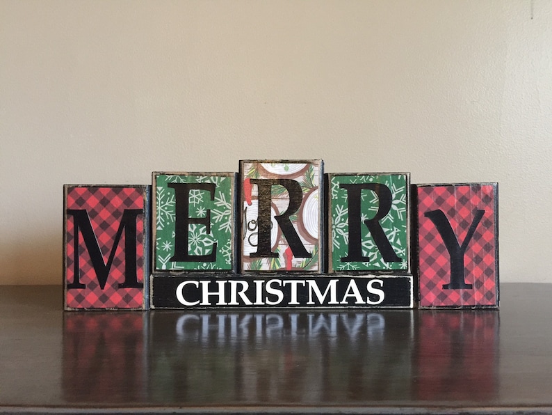 May include: Wooden block letters spelling "Merry Christmas" with red and green plaid patterns and a black background. The letters are arranged in a line on a dark brown surface.