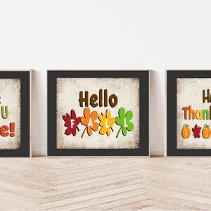 May include: Three framed prints with Thanksgiving-themed designs. The first print has a black background with white text that reads "Gobble Till You Wobble!" The second print has a black background with white text that reads "Hello Fall" and features five colorful leaves. The third print has a black background with white text that reads "Happy Thanksgiving" and features five orange pumpkins and five red, orange, and yellow leaves.