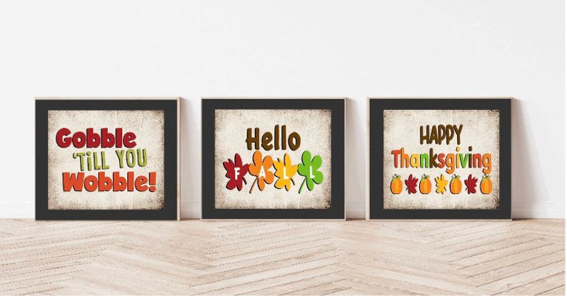 May include: Three framed prints with Thanksgiving-themed designs. The first print has a black background with white text that reads "Gobble Till You Wobble!" The second print has a black background with white text that reads "Hello Fall" and features five colorful leaves. The third print has a black background with white text that reads "Happy Thanksgiving" and features five orange pumpkins and five red, orange, and yellow leaves.