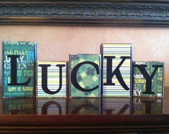 May include: Wooden blocks spelling out the word "LUCKY" in black letters. Each block is covered in green, white, and yellow patterned paper with shamrock designs.