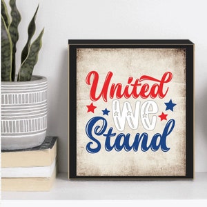 以下が含まれることがあります： 赤、白、青で「United We Stand」という言葉が書かれた額入りのプリント。デザインには星とヴィンテージスタイルの背景が含まれています。アートワークは白い表面に表示されています。