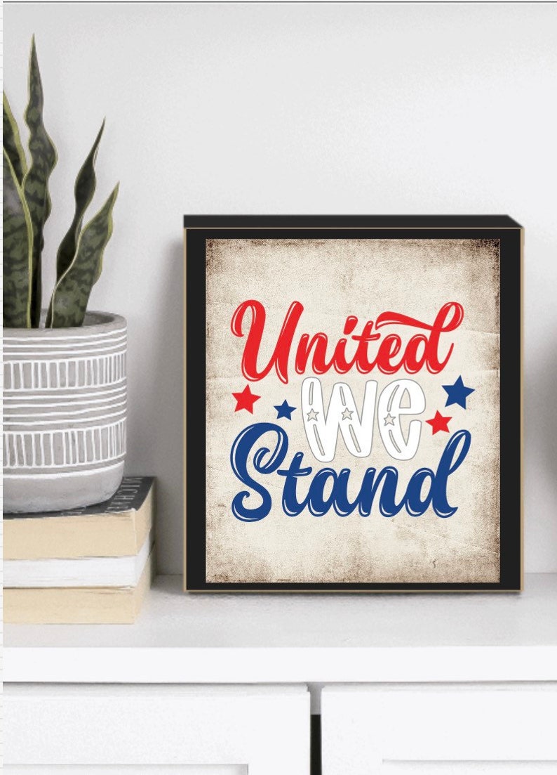 以下が含まれることがあります： 赤、白、青で「United We Stand」という言葉が書かれた額入りのプリント。デザインには星とヴィンテージスタイルの背景が含まれています。アートワークは白い表面に表示されています。