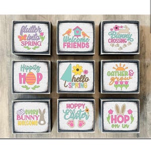 May include: Nine wooden signs with various Easter-themed sayings and graphics. The signs are painted white with black borders and feature colorful graphics and text. The sayings include "Flutter into Spring", "Welcome Friends", "Bunny Crossing", "Hippity Hop", "Hello Spring", "Gather & Grow", "Every Bunny Welcome", "Hoppy Easter", and "Hop On In".