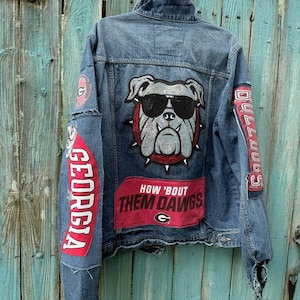 May include: A blue denim jacket featuring a large patch of a bulldog wearing sunglasses on the back. The jacket includes red patches with the text GEORGIA, BULLDOGS, and HOW 'BOUT THEM DAWGS, along with a circular G logo on the upper sleeve.