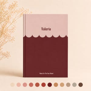 May include: A hardcover notebook with a two-tone cover featuring a light pink top section and a dark maroon bottom section separated by a scalloped line. The name Valeria is printed in dark text, with the words Notes For The Year Ahead below.