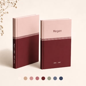May include: Two hardcover planners with a two-tone design featuring a light pink upper section and a dark maroon lower section separated by thin horizontal lines. The front cover displays the name Megan and the years 2026-2027 in dark text.