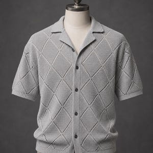 May include: A light gray short-sleeved knit shirt featuring a textured diamond pattern throughout the fabric. The garment has a collared neckline, a button-down front with four dark buttons, and ribbed trim along the sleeves and the bottom hem.