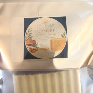 Op de afbeelding: Een rechthoekig stuk gele kurkumazeep verpakt in een goudkleurig metallic zakje met een doorzichtig venster. Het etiket bevat de tekst Ethereal JD, Turmeric Glow Soap en afbeeldingen van kurkumawortel en poeder, met een lotusbloem op de zeep.