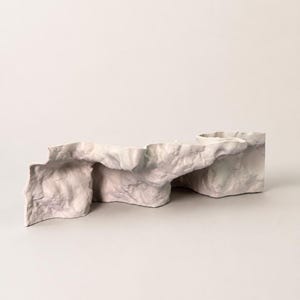 Puede incluir: Un objeto escult&oacute;rico de forma irregular y ondulada con una superficie mate de color blanco roto. La pieza presenta grietas profundas, crestas y una textura similar a la roca con sutiles sombras gris&aacute;ceas y p&uacute;rpuras sobre su forma org&aacute;nica.