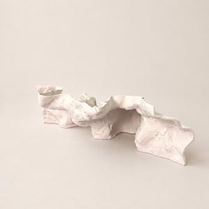 Puede incluir: Una escultura de cer&aacute;mica de color rosa p&aacute;lido con una forma larga, ondulada y arrugada. El objeto presenta una superficie mate y texturizada con curvas org&aacute;nicas y fluidas que crean pliegues y sombras profundas, similar a una tela drapeada.