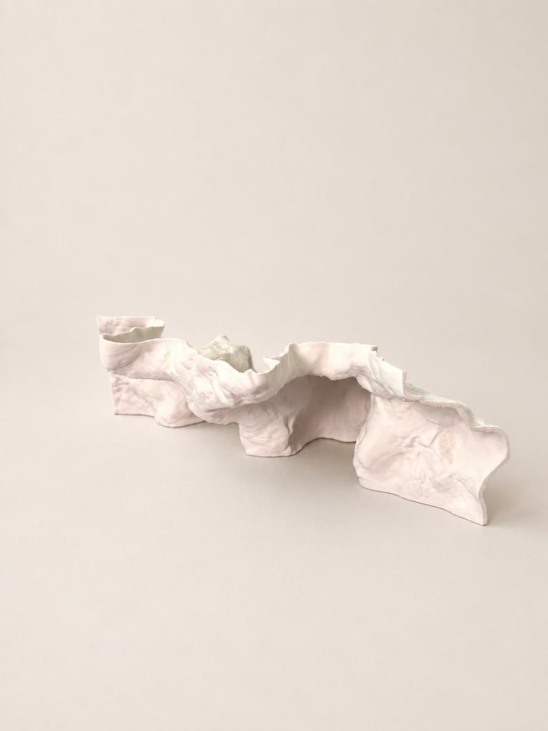 Puede incluir: Una escultura de cer&aacute;mica de color rosa p&aacute;lido con una forma larga, ondulada y arrugada. El objeto presenta una superficie mate y texturizada con curvas org&aacute;nicas y fluidas que crean pliegues y sombras profundas, similar a una tela drapeada.